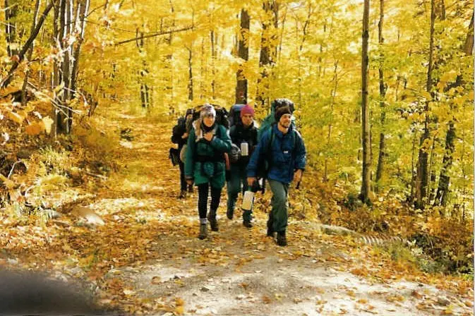 three friends with backpacks hiking a trail through yellow fall woods